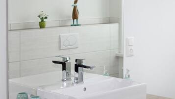 A modern bathroom with a white sink, two faucets, and a mirror above. There is a small plant on the shelf above the mirror and a soap dispenser on the wall.