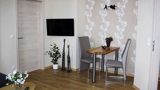 Una sala de estar con piso de madera, un televisor montado en la pared, una mesa de comedor con sillas y una pequeña planta sobre la mesa.