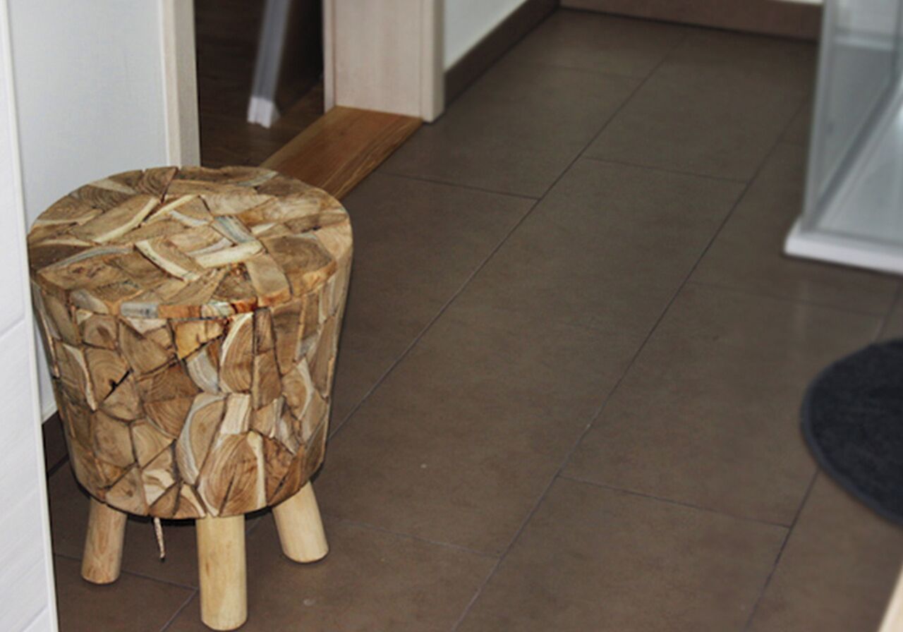 Un tabouret rond en bois, orné d’un motif tissé, posé sur un sol carrelé dans un couloir.