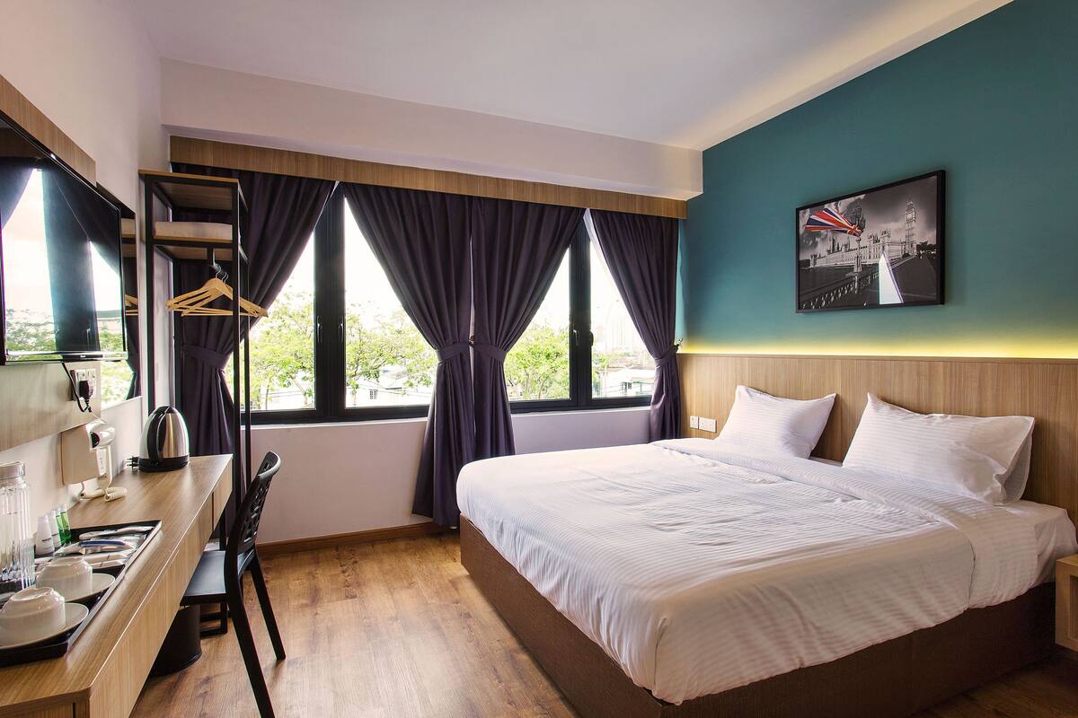 Habitación de hotel con cama, escritorio con hervidor, silla y ventana con cortinas.
