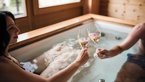 A person in a bathtub with another person outside the tub, holding champagne glasses.