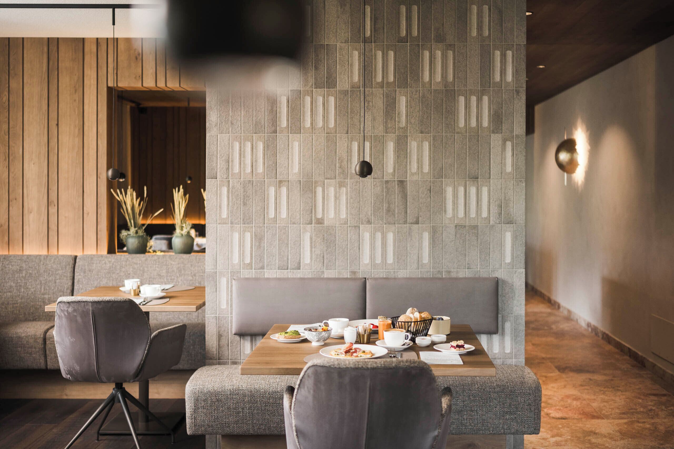 A modern dining area with a wooden wall, grey cushioned seating, and a table set with breakfast.