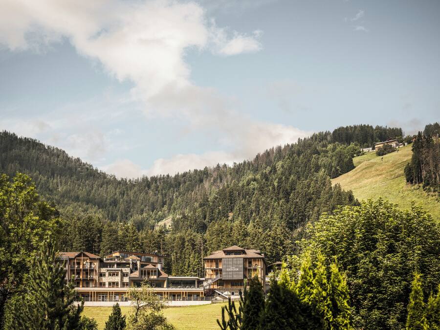 A multi-story lodge with a modern architectural style, surrounded by dense forest and hills.
