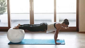 A person performing a yoga exercise on a blue mat with a large white exercise ball, in a room with large windows and wooden flooring.