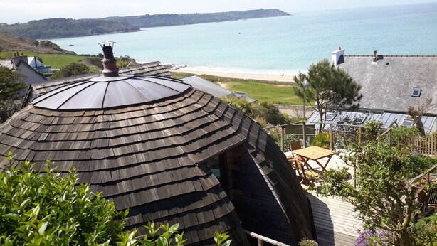 A coastal property with a wooden gabled roof, a patio area with outdoor furniture, and a view of the sea and beach. - Original round wood house T3 sea view (Locquirec)