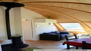A cozy loft-style room with a high ceiling, wooden beams, a built-in fireplace, a sofa, and a dining table. - Original round wood house T3 sea view (Locquirec)