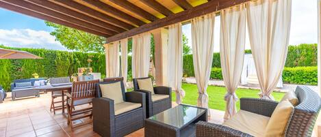 Eine Terrasse im Freien mit Korbmöbeln, einem Tisch mit Glasplatte und Blick auf einen gepflegten Garten.