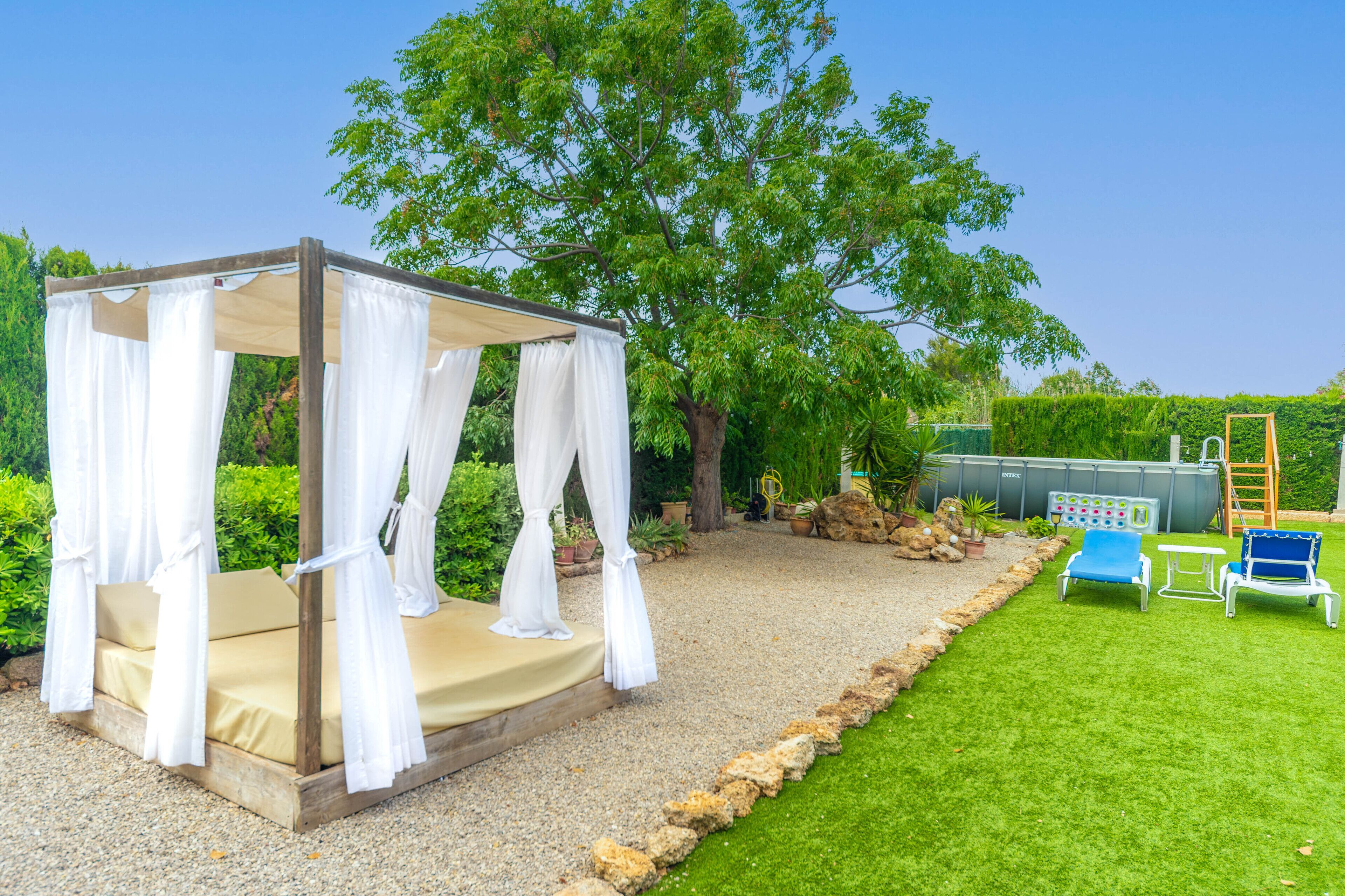 A gazebo with a beige floor and white curtains, surrounded by a gravel path and green grass.