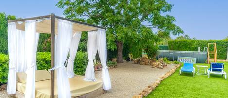 A gazebo with a beige floor and white curtains, surrounded by a gravel path and green grass.