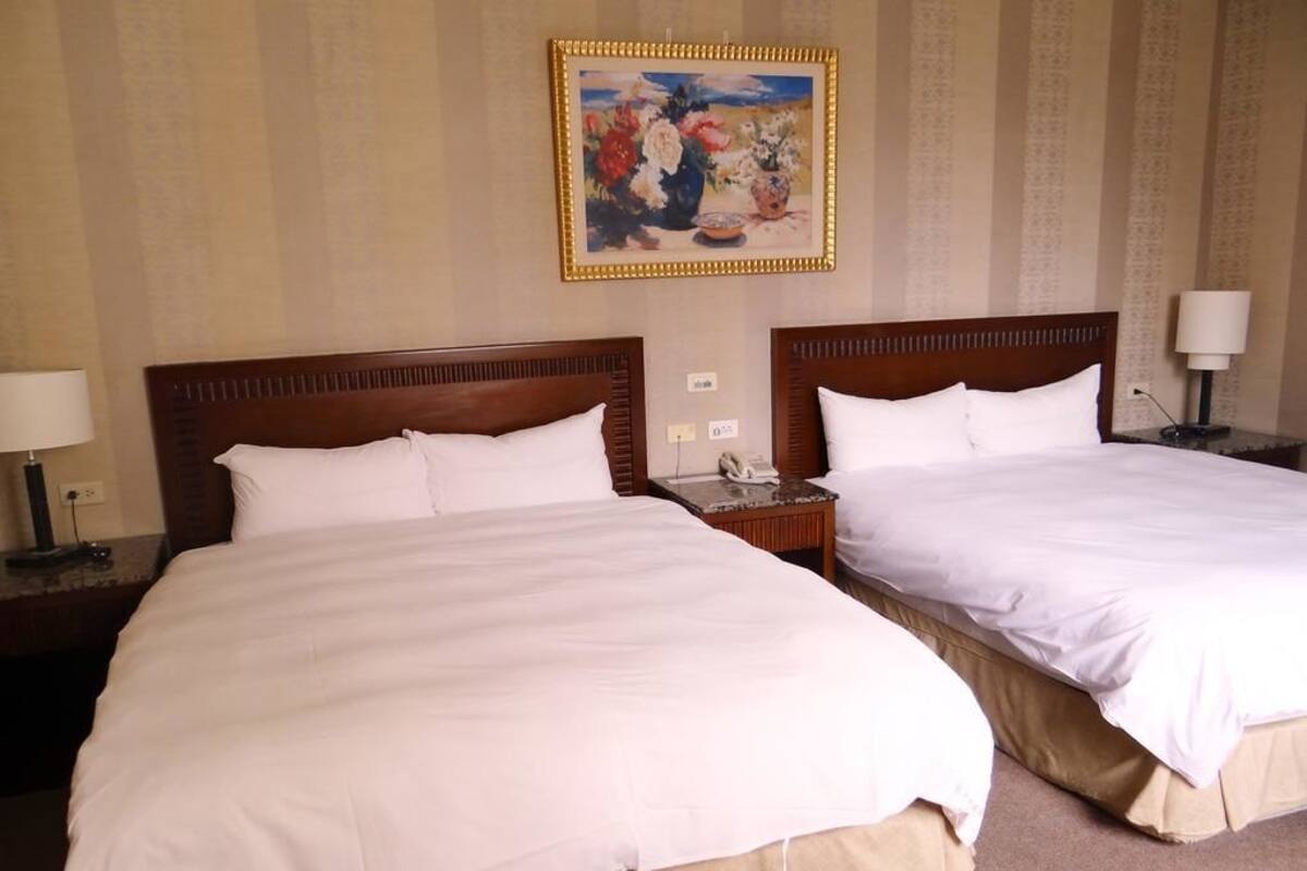 A hotel room with two beds, a painting on the wall, and a nightstand with a lamp.