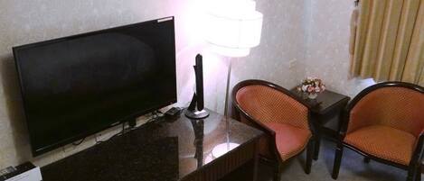 A hotel room with a flat-screen TV, a desk with a lamp, two orange chairs, and a small table with a decorative item.