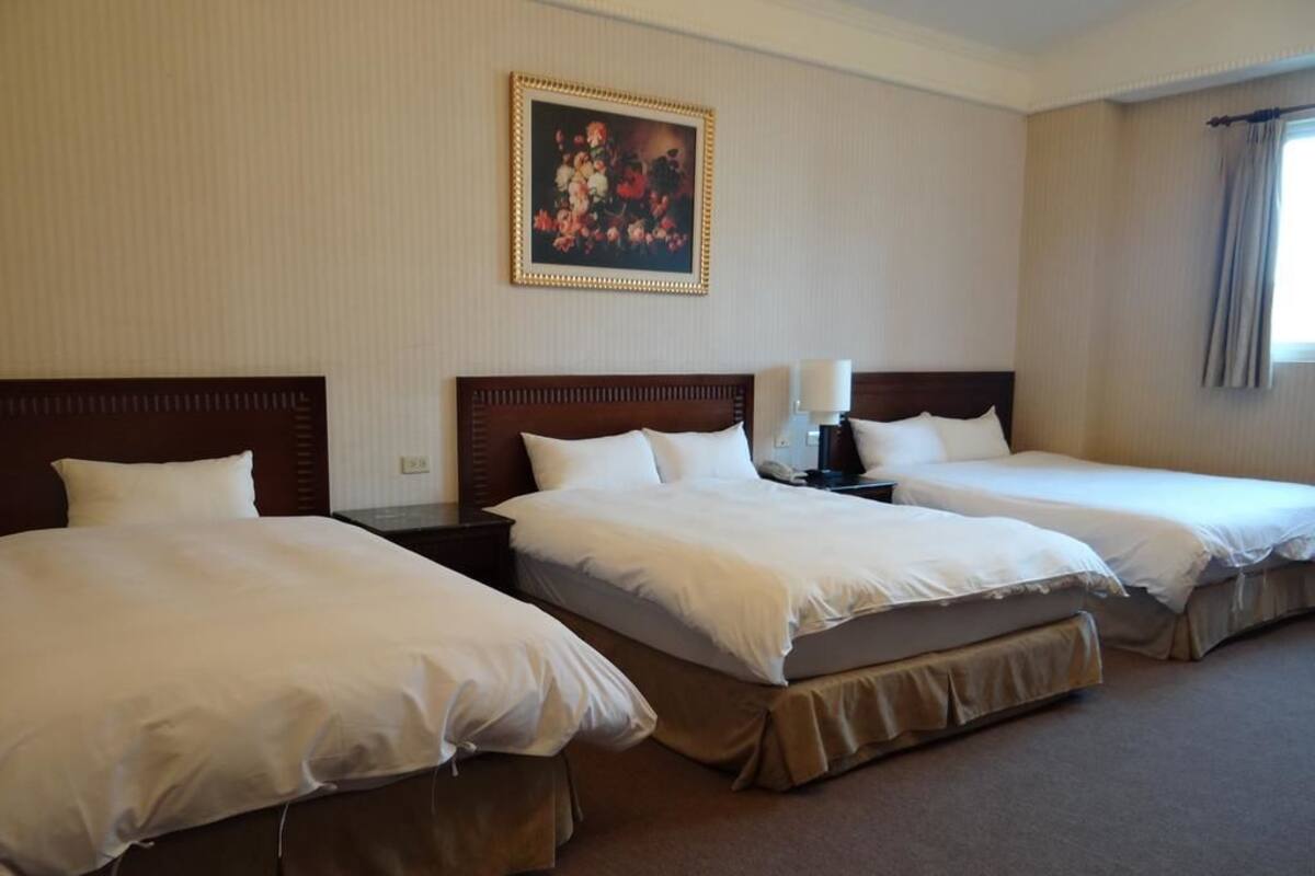 A hotel room with three beds, a painting on the wall, and a window with curtains.