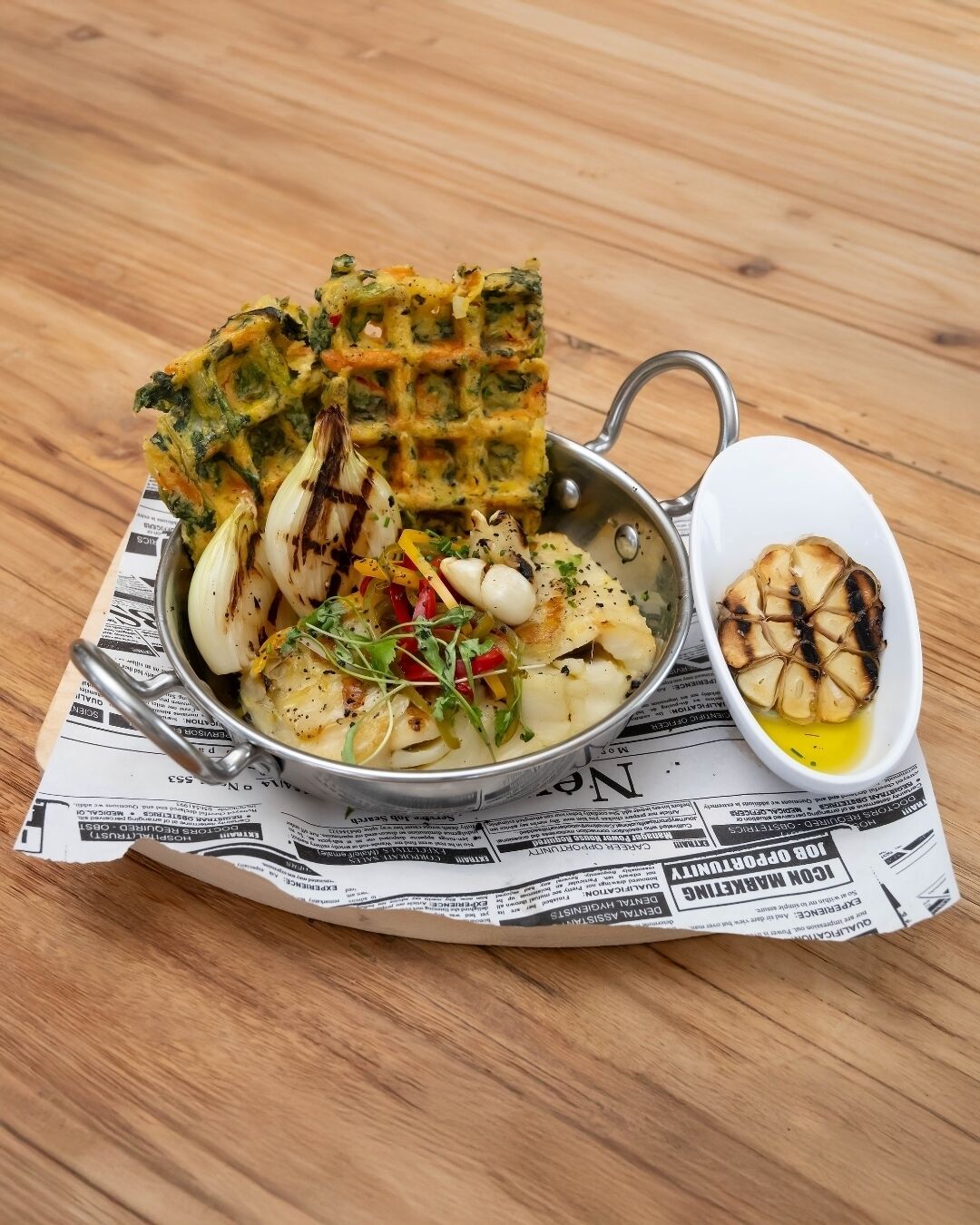 A meal consisting of grilled vegetables, a savory pancake, and a bowl of garlic cloves on a wooden table.