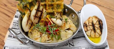 A meal consisting of grilled vegetables, a savory pancake, and a bowl of garlic cloves on a wooden table.