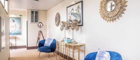 A hallway with blue upholstered chairs, a gold-framed mirror, and artwork on the walls.