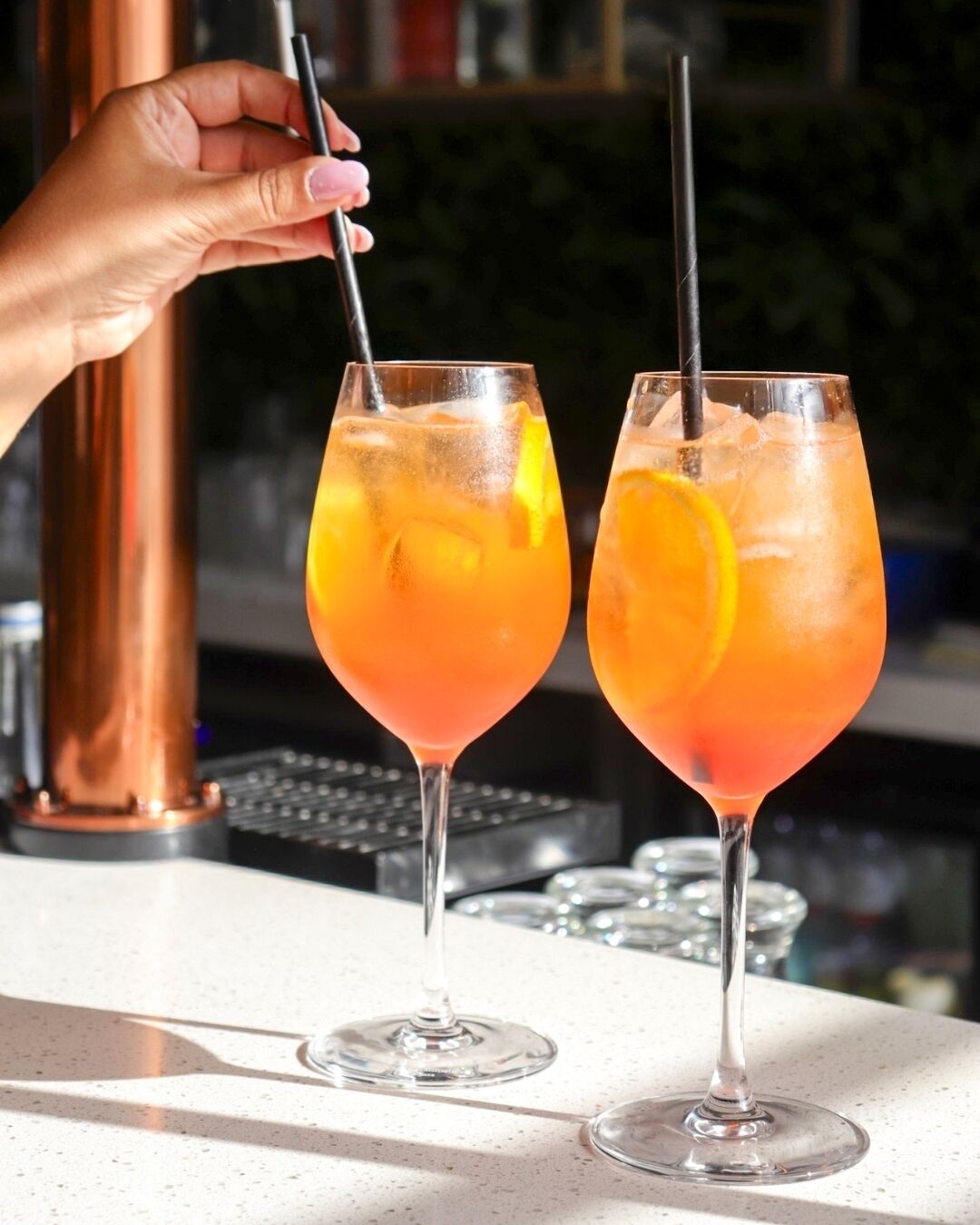 Two glasses of a pinkish-orange cocktail with ice and a slice of orange, served in stemmed glasses with straws, on a bar counter.