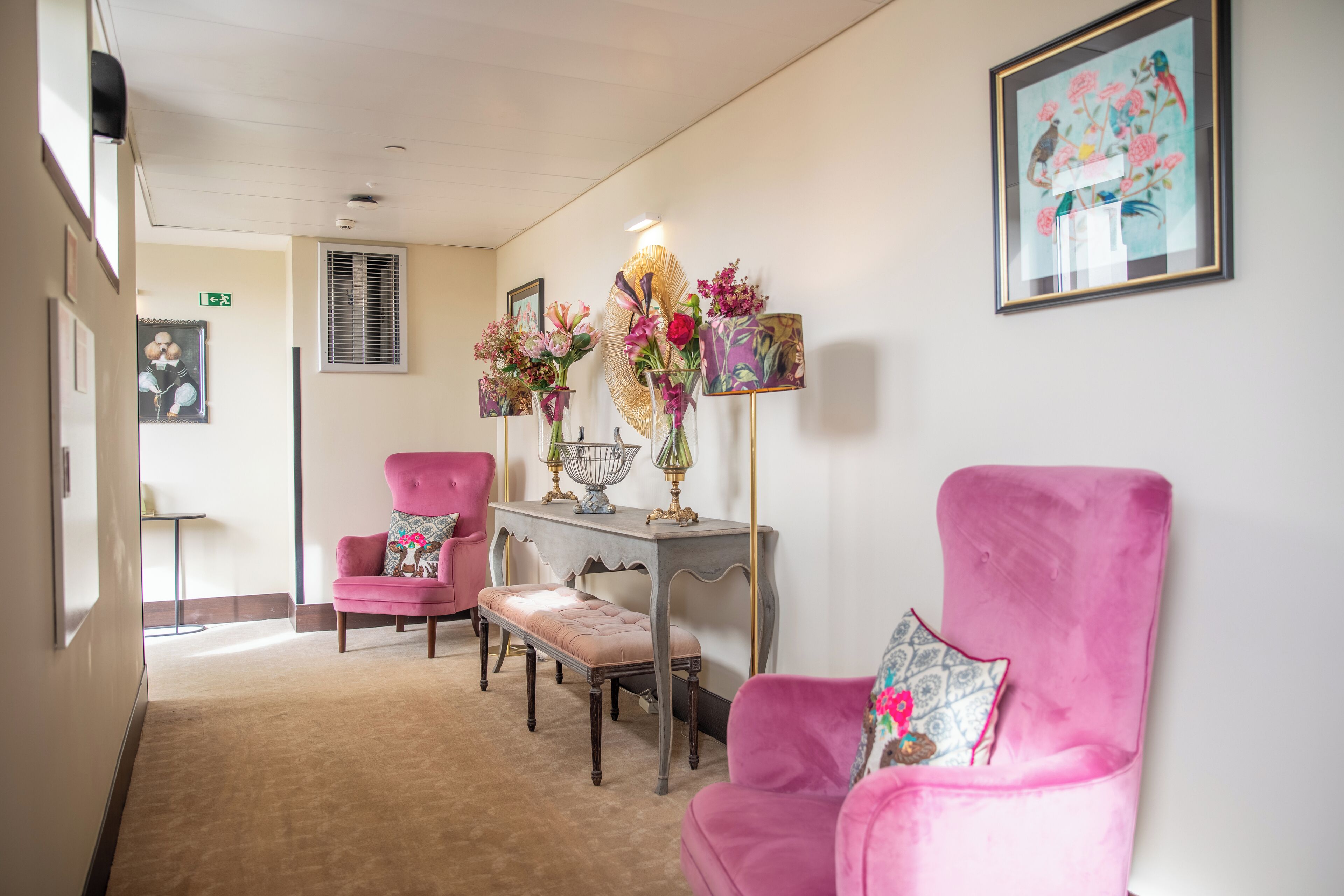 A hallway with two pink armchairs, a bench, and a table with floral arrangements.