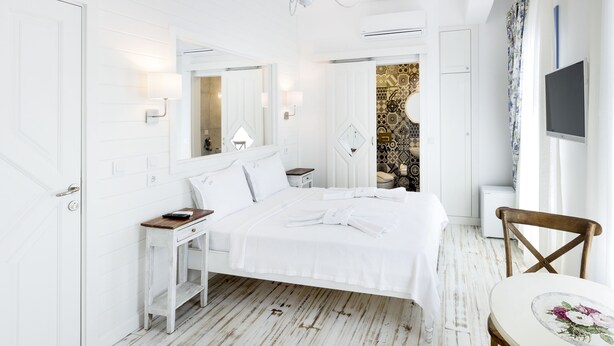 A white-themed bedroom with a bed, bedside table, and a small round table with a floral tablecloth. - Bademlik Otel (Bozcaada)