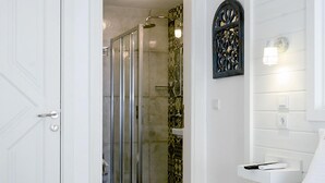 A hotel room with a bed, a bedside table, a nightstand, a shower area, and a door. - Bademlik Otel (Bozcaada)