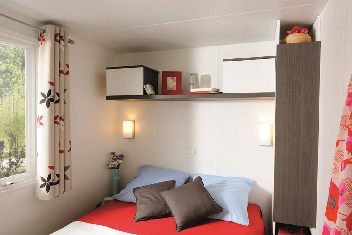 Een slaapkamer met een bed, kussens, een nachtkastje, een plank met boeken en een fotolijst, en een raam met gordijnen.