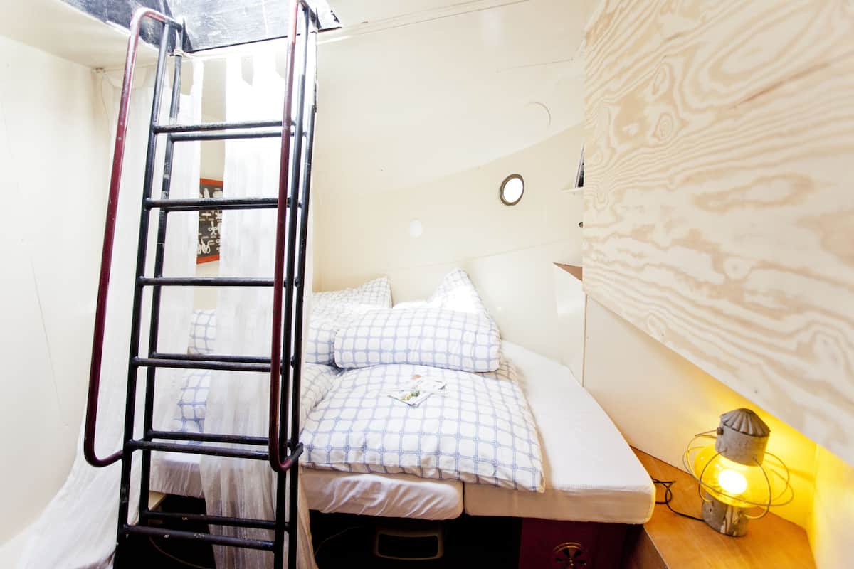 A small, compact room with a bunk bed, a ladder, a bedside table with a lamp, and a wall with a wood-like pattern.