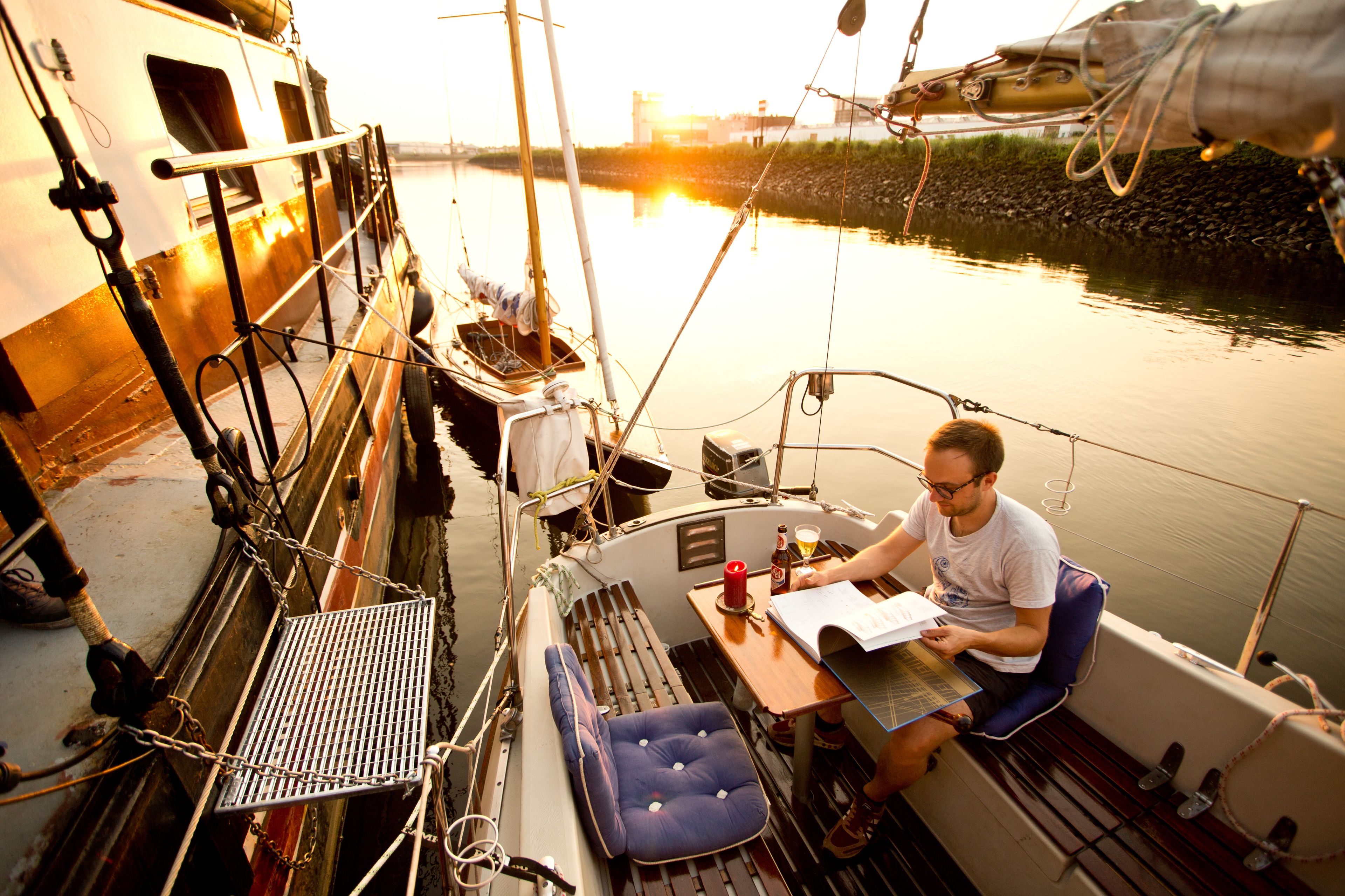 Ein Boot mit Tisch und Stuhl, eine Person liest ein Buch, im Hintergrund liegt ruhiges Wasser.