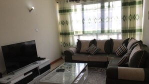 A living room with a flat-screen TV, a glass coffee table, a sofa set, and a patterned curtain. - Jambo Homestay (Syokimau)
