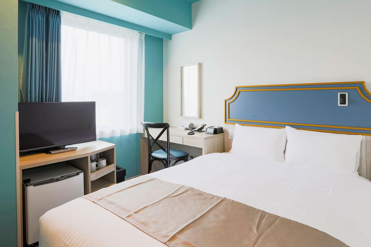 Hotellihuone, jossa on sänky, työpöytä, televisio, tuoli ja ikkuna, jossa on verhot.