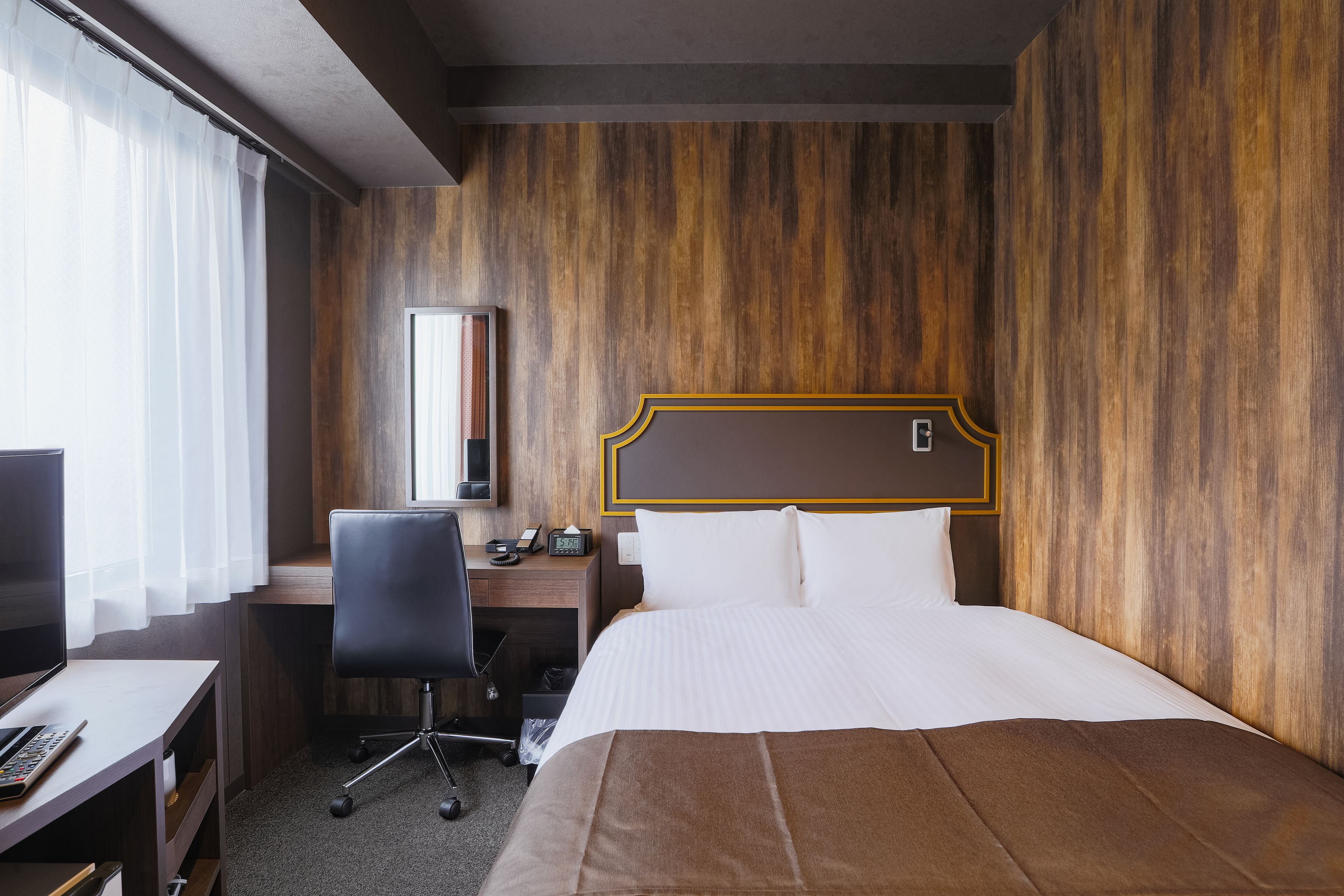 Habitación de hotel con pared de madera, cama con ropa blanca, escritorio con computadora y silla, y televisor.