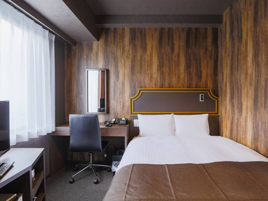 Habitación de hotel con pared de madera, cama con ropa blanca, escritorio con computadora y silla, y televisor.