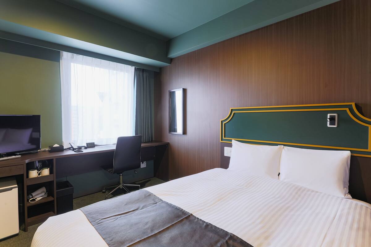 Hotellihuone, jossa on sänky, työpöytä, televisio ja ikkuna, jossa on verhot.
