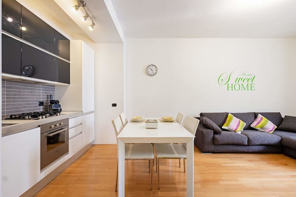 A modern kitchen with a white table and grey sofa, a clock, and a "Home Sweet Home" sign.