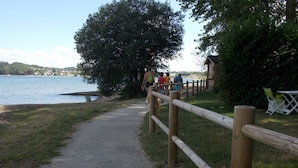 A gravel path leading to a wooden fence by a lake, with a small building and green chairs in the background.