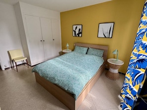 Un dormitorio con una cama, dos cuadros en la pared, una silla, una mesita de noche y una cortina.
