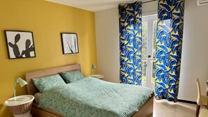 A bedroom with a bed, two wall art pieces, a window with curtains, a bedside table, a lamp, and a chair.