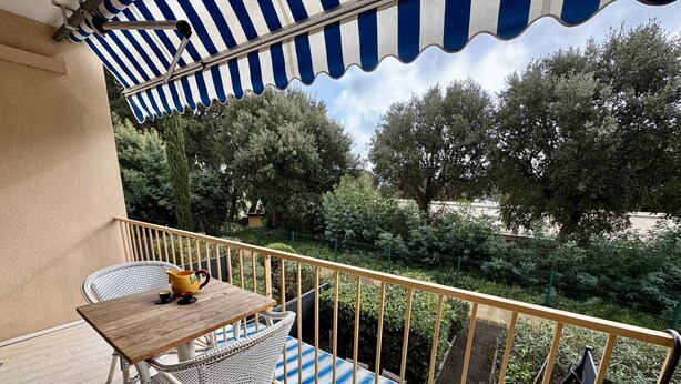 A balcony with a striped awning, a wooden table, wicker chairs, and a view of trees and greenery.