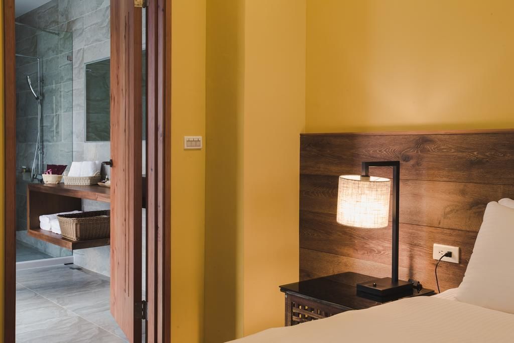 A hotel room with a wooden headboard, a bedside lamp, a bed, and a bathroom with a shower and a sink.