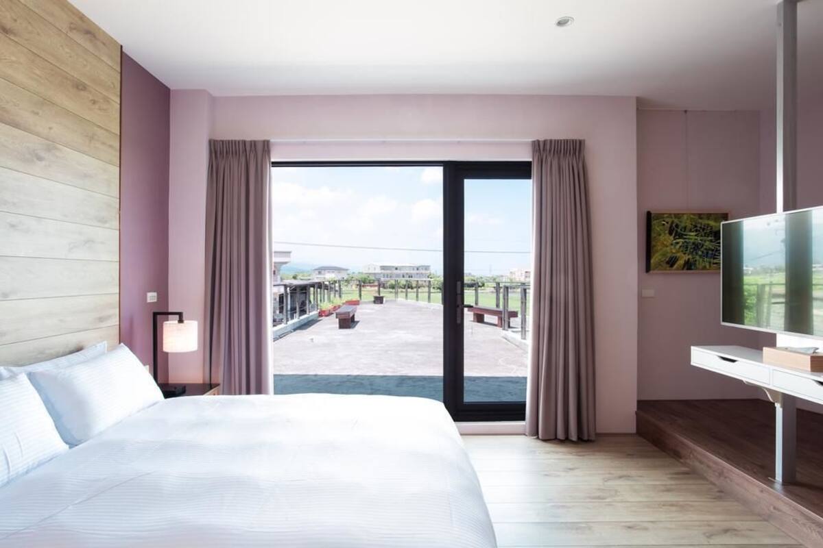 A hotel room with a large bed, a desk with a TV, and a balcony with outdoor seating.