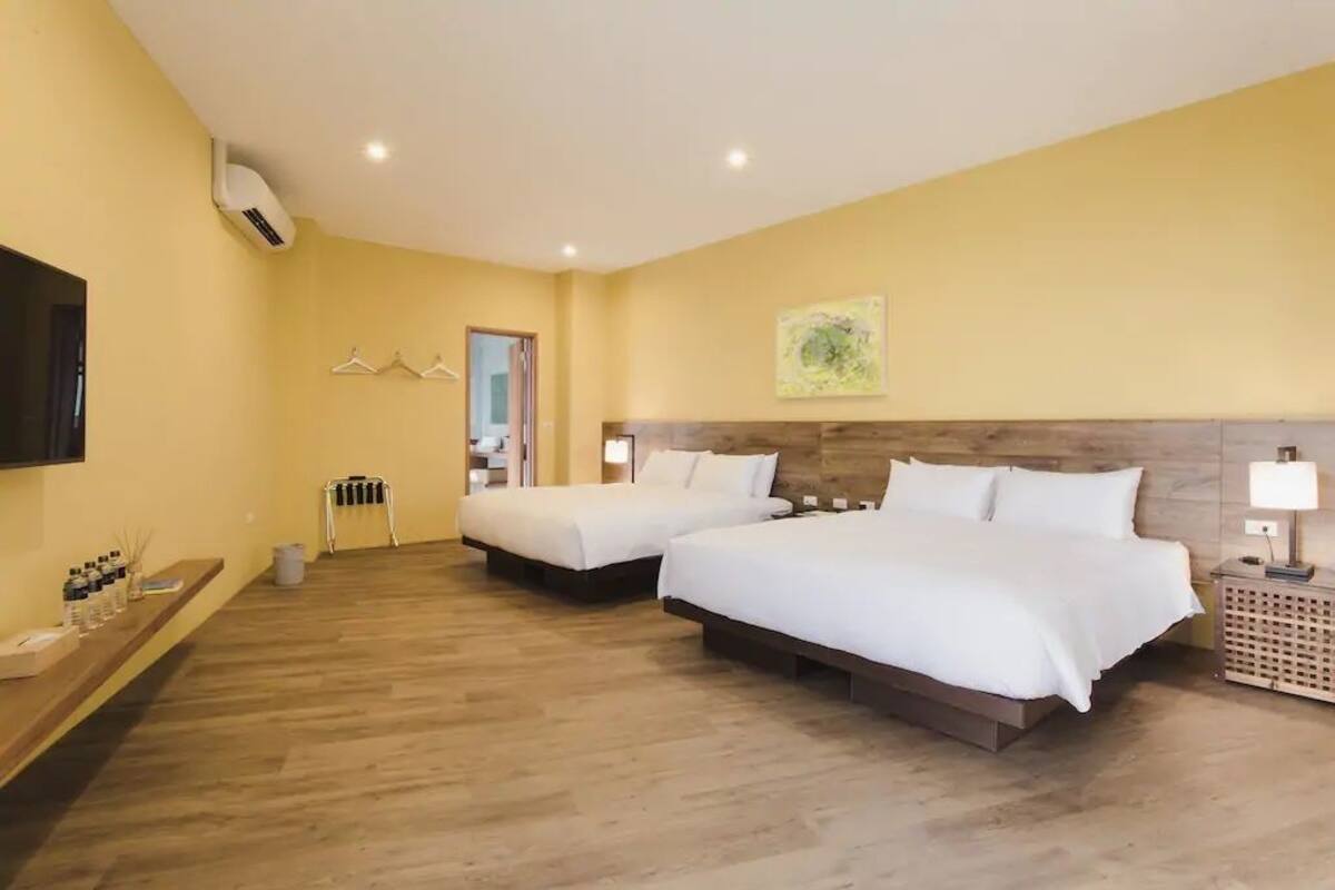 A hotel room with two beds, a wooden floor, a mounted TV, and a wall-mounted air conditioner.