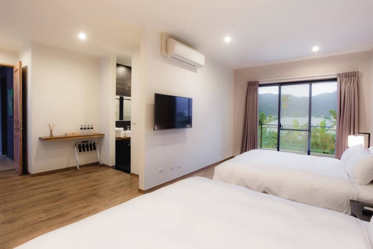 A hotel room with two beds, a TV, an air conditioner, a desk with a chair, and a window with a view of mountains and a lake.
