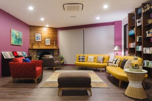 Una sala de estar moderna con un sillón rojo, un sofá amarillo y un reposapiés marrón. Se observa una estantería llena de libros y objetos decorativos.