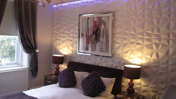 Une chambre à coucher avec un lit, deux tables de chevet, un lustre et un mur orné d’un motif géométrique.