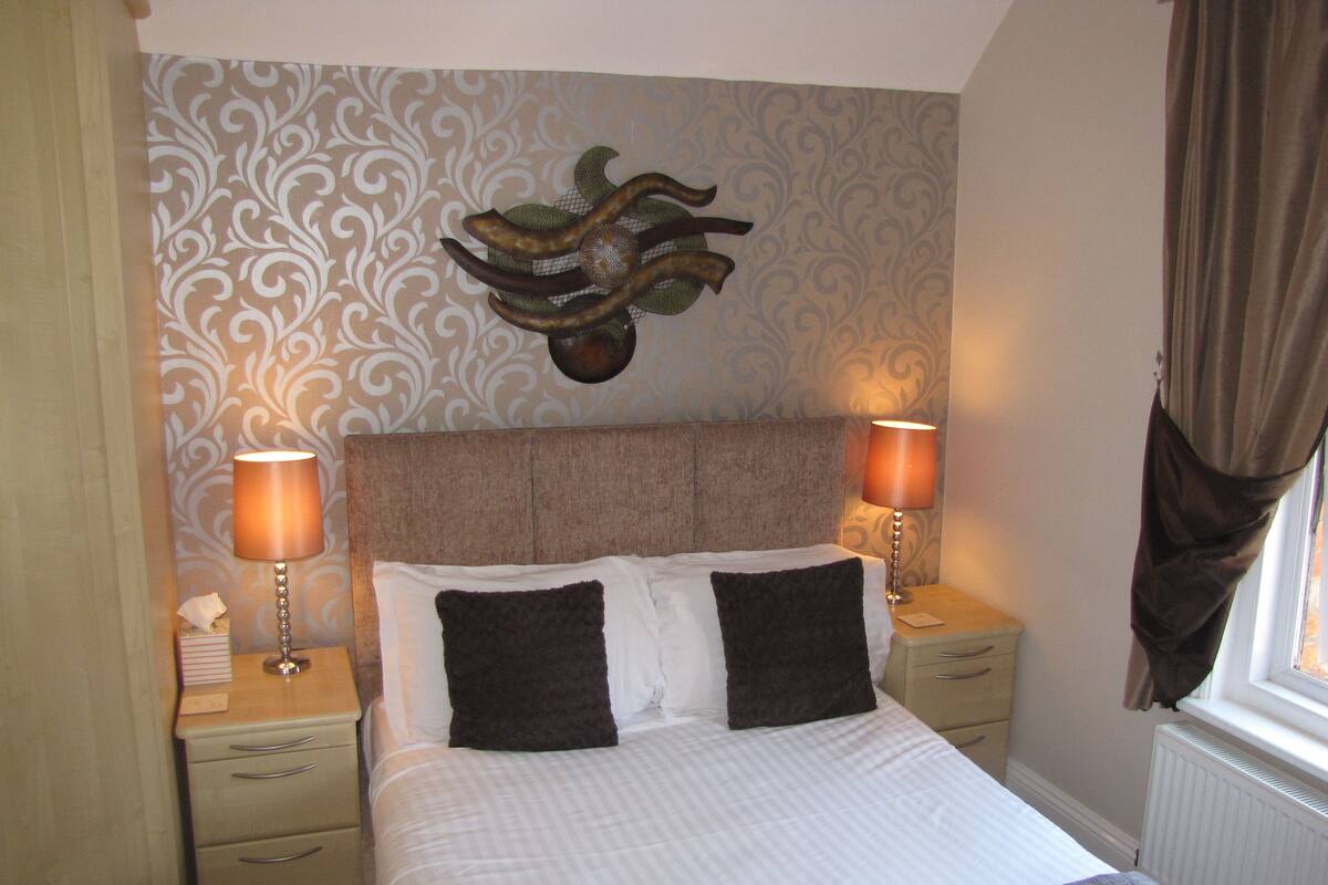 A neatly made bed with two pillows, two bedside lamps, a decorative wall piece, and a window with curtains.