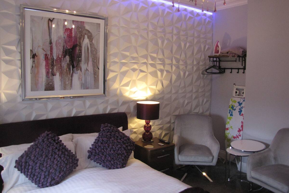 A modern hotel room with a geometric-patterned wall, a framed artwork, a bed with purple pillows, a bedside lamp, a grey armchair, and a small round table.
