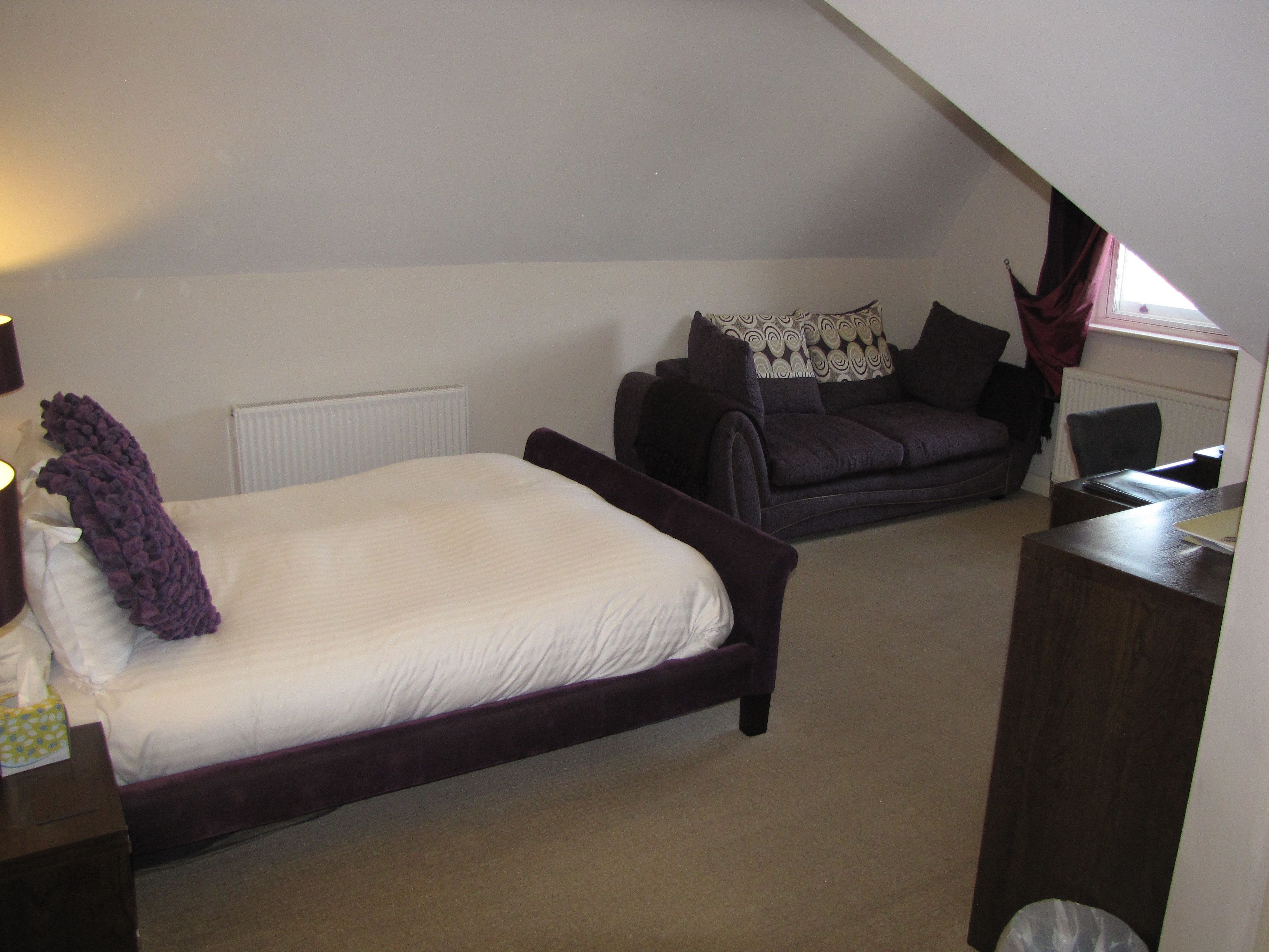 Ein Schlafzimmer mit einem Bett, einem dunkelgrauen Sofa, einem kleinen Tisch und einem Fenster mit Vorhängen.