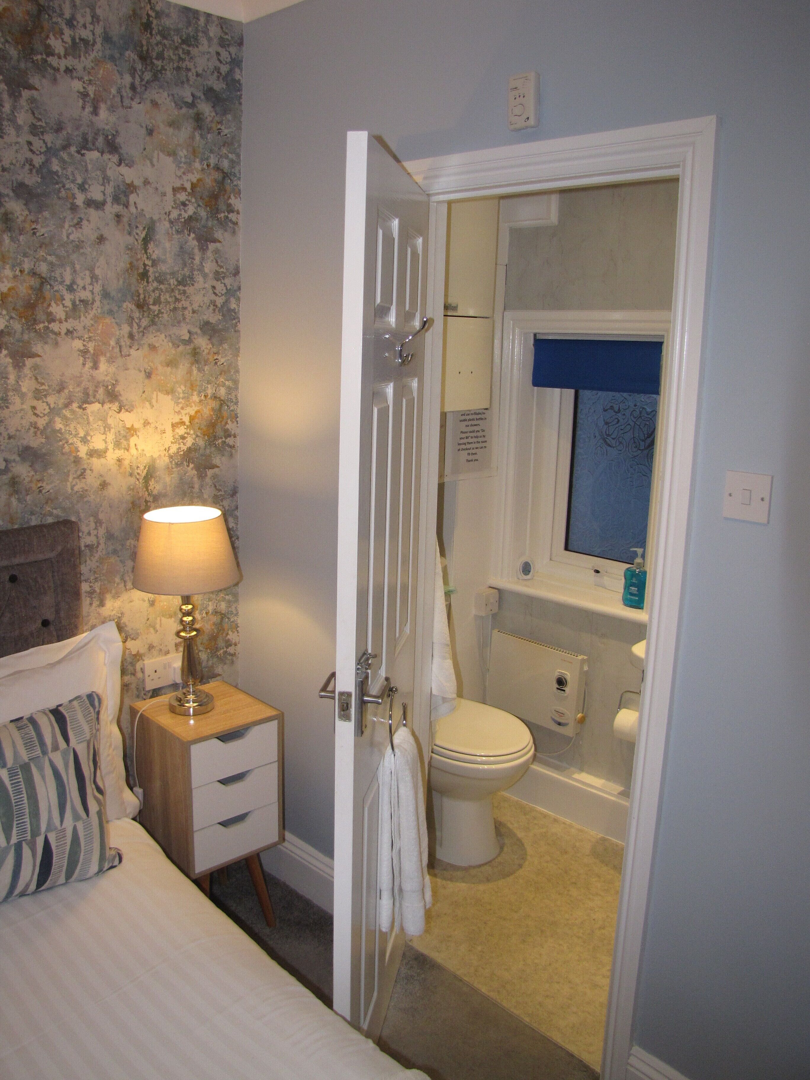 A bedroom with a bed, bedside table, lamp, and a bathroom visible through an open door.