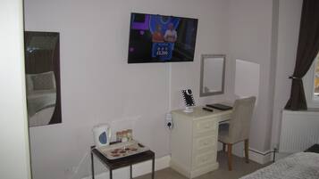 Una habitación de hotel con cama, escritorio, silla, televisor, espejo, una mesita con hervidor y radiador.