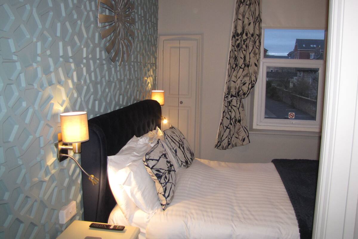A bedroom with a patterned wall, a bed with white bedding, two bedside lamps, and a window with a view of buildings.