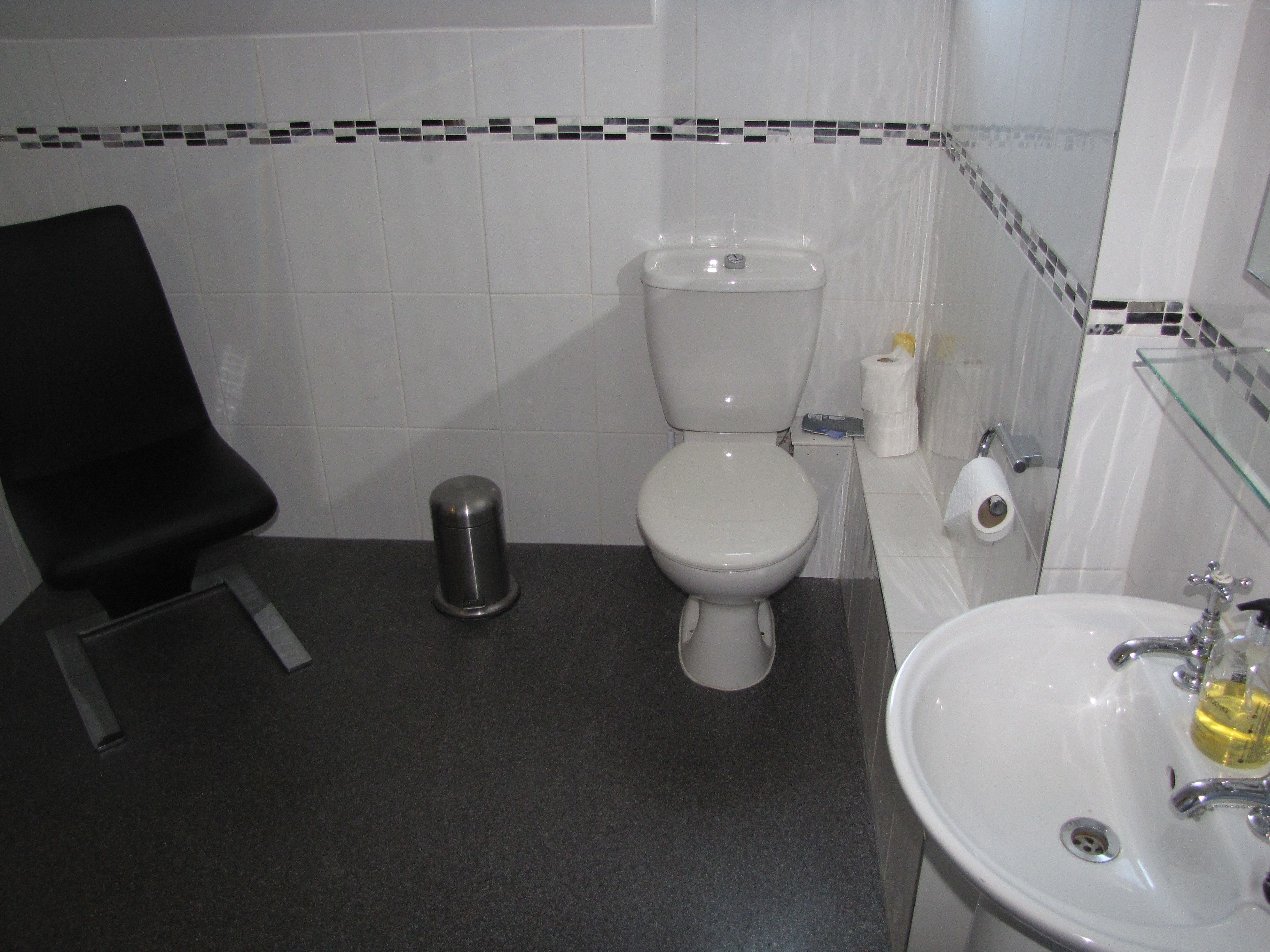 A bathroom with a toilet, a trash can, a sink, and a black chair.