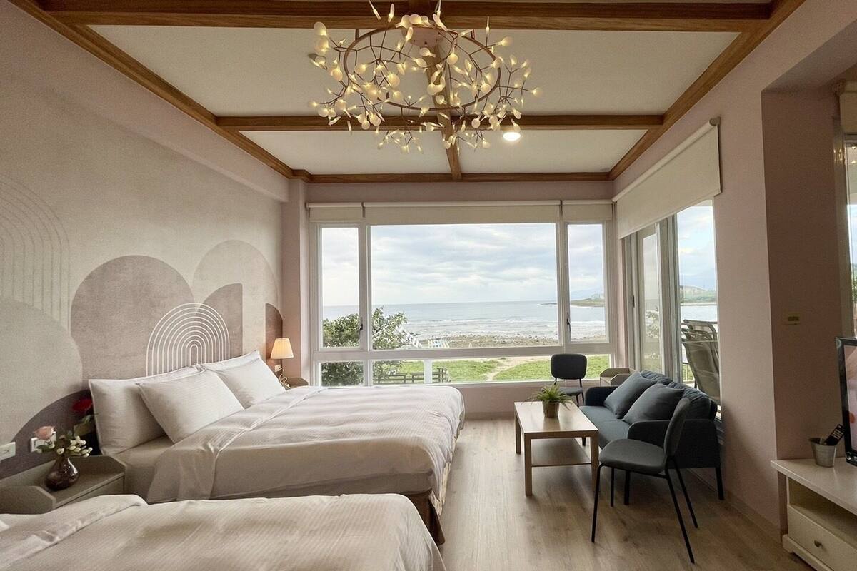 Habitación de hotel con dos camas, un sofá, un televisor y ventanas amplias con vista al mar.